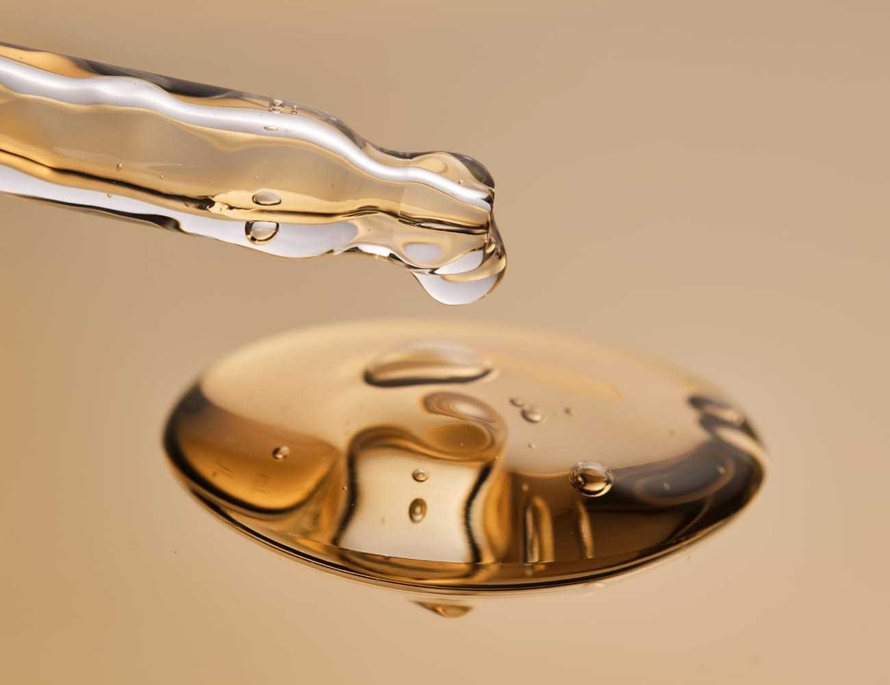 A dropper releasing oil into a puddle against a beige background.